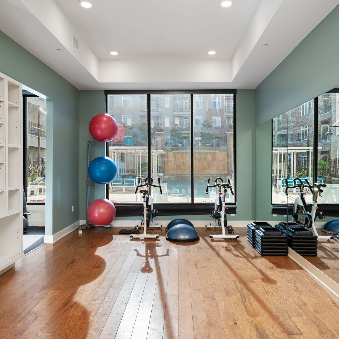 Domain City Center A well-lit home gym with wooden flooring, featuring an exercise bike, stability balls stacked in a corner, a BOSU ball, a set of stackable aerobic steps, and floor-to-ceiling mirrors.